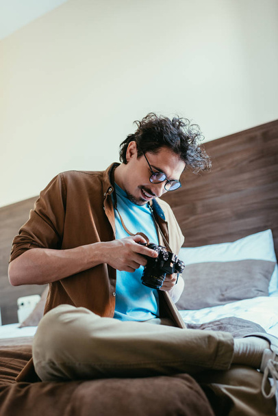 Erwachsener Mann mit Brille blickt auf Fotokamera und sitzt auf Bett im Hotelzimmer - Foto, Bild