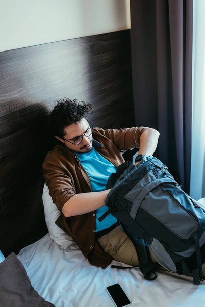 turista masculino tomando algo de la mochila mientras está sentado en la cama con teléfono inteligente en el hotel
  - Foto, imagen