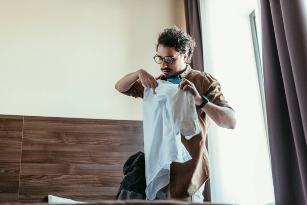 male tourist in eyeglasses putting shirt into backpack in hotel room - Foto, afbeelding