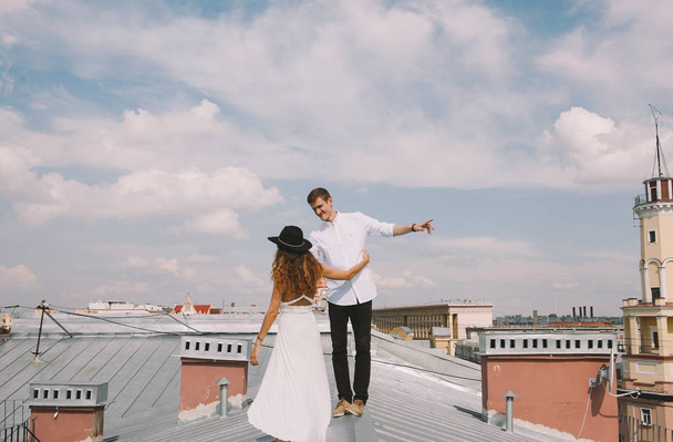 loving couple - a girl in a white dress and a hat and a guy - on a walk on the roof hugging and laughing, a view from the height of the city, positive and sincere emotions of love - Foto, Imagem