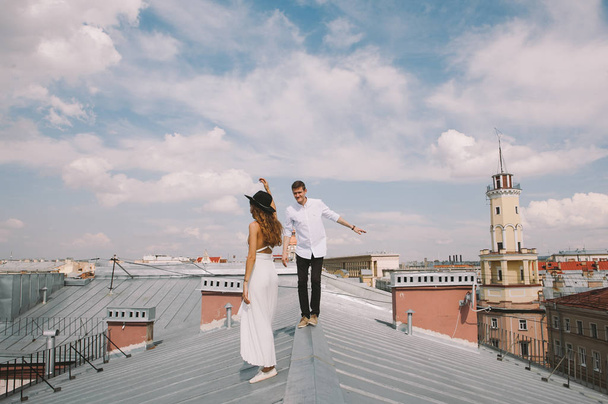 loving couple - a girl in a white dress and a hat and a guy - on a walk on the roof hugging and laughing, a view from the height of the city, positive and sincere emotions of love - Φωτογραφία, εικόνα