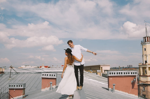 loving couple - a girl in a white dress and a hat and a guy - on a walk on the roof hugging and laughing, a view from the height of the city, positive and sincere emotions of love - Valokuva, kuva
