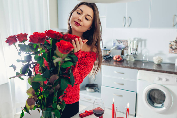 Jonge vrouw gevonden boeket rozen met kaars, wijn en cadeau doos op de keuken. Gelukkig meisje ruikende bloemen. Verrassing van de dag van Valentijnskaarten - Foto, afbeelding