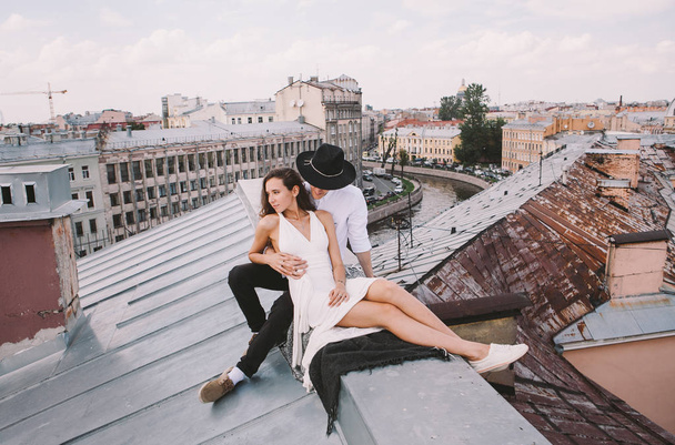 loving couple - a girl in a white dress and a guy with a hat - on a walk on the roof hugging and laughing, a view from the roof of the city, positive and sincere emotions of love - Фото, изображение