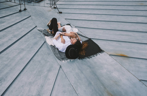 couple in love lying on the roof of the house and hugging - a girl in a white dress and a guy with a hat - view from the roof of the city, positive and sincere emotions of love - Foto, Imagen
