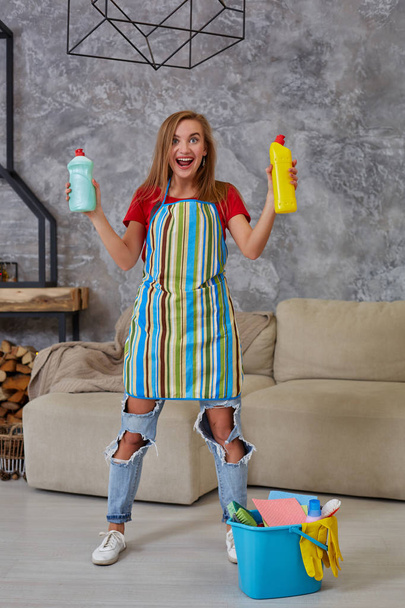 Kadın karşılaştırır iki deterjan şişesi evde oturma odasında mavi kova içinde birçok ev objeleri temizlik. Ev iş. - Fotoğraf, Görsel