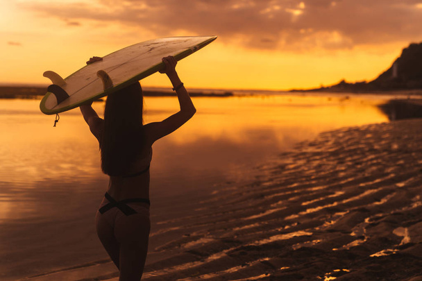 Silhouette of slim girl with surfboard in hands at beach on background of beautiful sunset - Fotografie, Obrázek