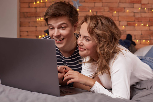 Sorrindo jovem casal homem e mulher deitado na cama e assistindo algo no laptop
 - Foto, Imagem