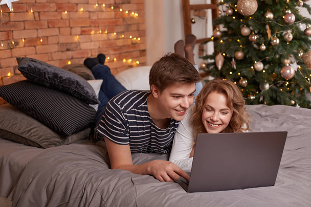 Lächelndes junges Paar Mann und Frau im Bett liegend und etwas auf dem Laptop beobachtend - Foto, Bild