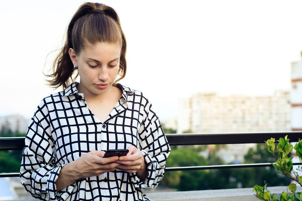 όμορφη νεαρή κοκκινομάλλα γυναίκα στέλνοντας μήνυμα με smartphone σε μια ταράτσα. - Φωτογραφία, εικόνα
