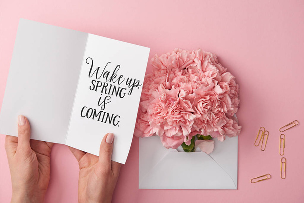 cropped view of woman holding greeting card with wake up, spring is coming lettering near carnation flowers in envelope on pink background - Photo, Image