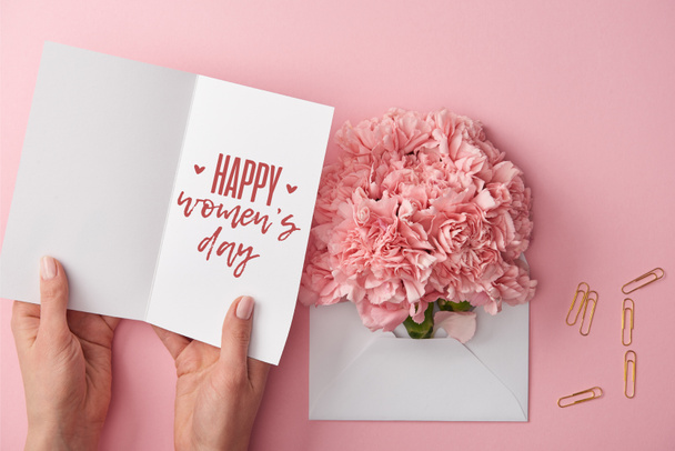 cropped view of woman holding greeting card with happy womens day lettering near carnation flowers in envelope on pink background - Foto, Imagem