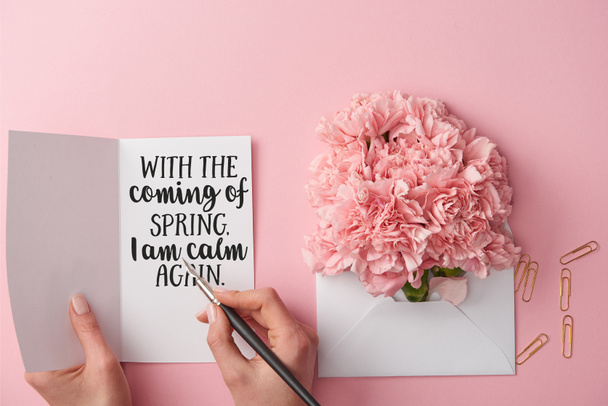 cropped view of woman holding spring greeting card and pen near carnation flowers in envelope on pink background - Photo, Image