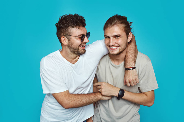 Portrait of two happy young hipster men embracing each other isolated over blue studio background - Fotoğraf, Görsel