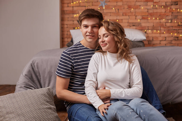 Portrait of a young loving couple resting on a grey couch together at home and hugging - Foto, Imagem