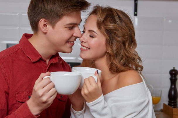 Χαρούμενος νεαρός υπέροχο ζευγάρι στην κουζίνα που αγκαλιάζονται μεταξύ τους. Απολαμβάνουν το να πίνεις καφέ - Φωτογραφία, εικόνα