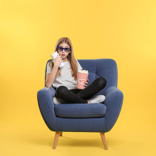 Emotional teenage girl with 3D glasses, popcorn and beverage sitting in armchair during cinema show on color background - Foto, Imagem