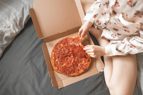 Una foto de cerca de una chica en pijama se sienta en la cama y toma un pedazo de pizza de la caja. Las piernas de la chica y una caja de pizza, toma un apetitoso pedazo de manos. Comer pizza en la cama
. - Foto, Imagen