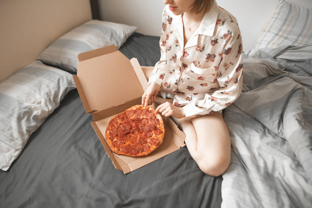 Pretty woman in a pajama sits on a bed with a pizza box and takes a piece. Girl sleeps pizza on the bed. Fast Food Concept. - Foto, immagini