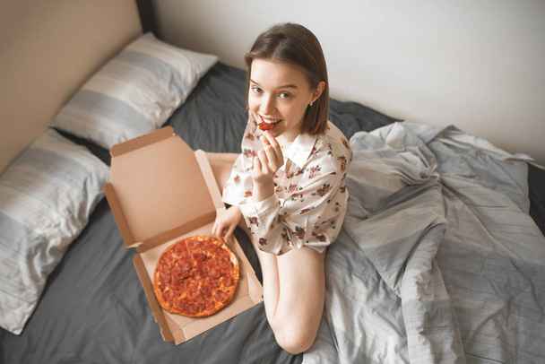 Portrait of an attractive girl in a pajama sitting on a bed with a box of pizza, biting a piece of pizza, looking into the camera and smiling. Happy woman eating pizza for breakfast. - Photo, Image