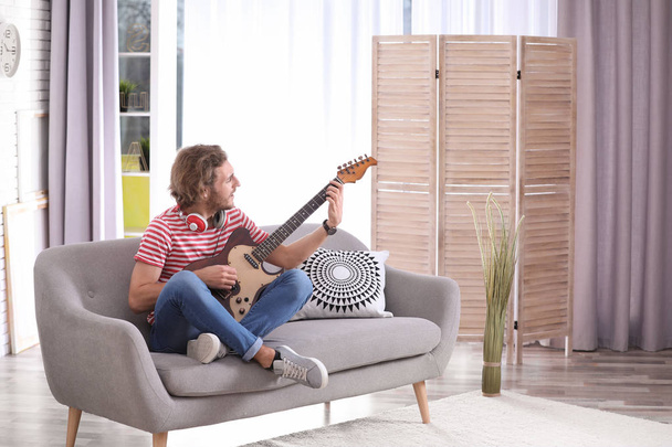 Young man with headphones playing electric guitar in living room. Space for text - 写真・画像