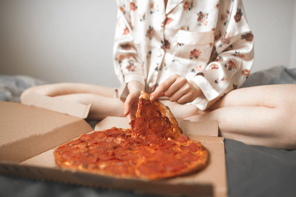 Girl is in pajamas sits at home on a bed and takes an appetizing piece of pizza from the box. Close up, Girl's hands take a delicious piece of pizza. Teenager is going to eat a piece of pizza. - Foto, Bild