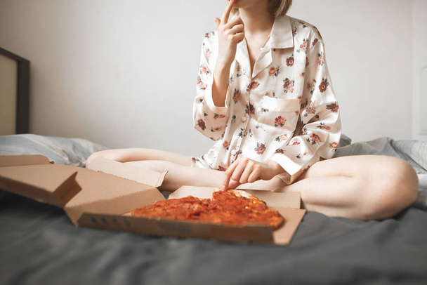 Meisje in een pyjama zit op het bed in de slaapkamer en een pizza eet uit het vak. Bijgesneden foto van een vrouw met een doos met pizza op het bed. Fastfood, Pizza levering. - Foto, afbeelding