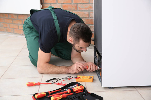 Male technician in uniform repairing refrigerator indoors - Foto, Imagen