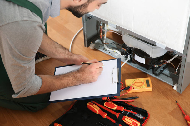 Male technician with clipboard examining broken refrigerator indoors, closeup - Foto, imagen