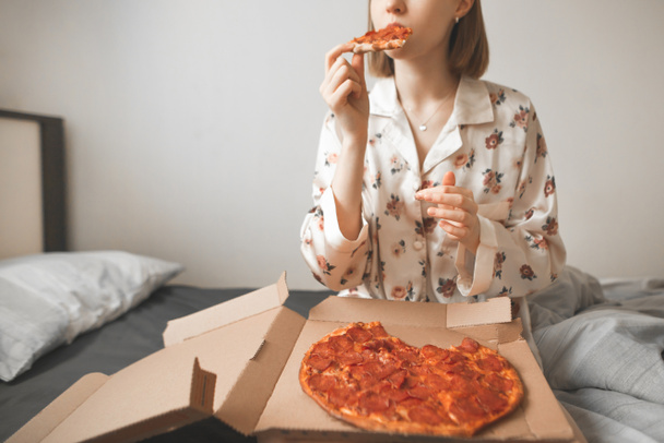 Csinos lány pizsama öltözött ült az ágy egy hangulatos hálószobából és pizzát eszik a listából, egy darab csípős és élvezem. Forró pizza az ágyban, a reggeli a napon ki. Pizza futár - Fotó, kép