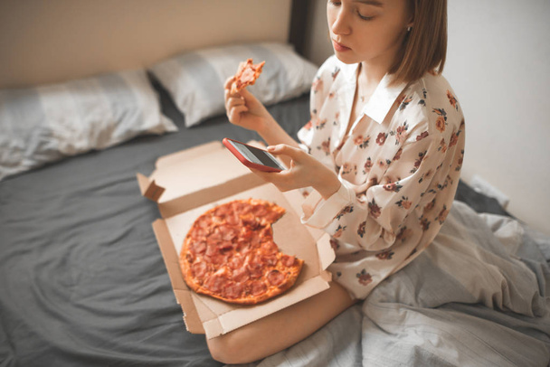 Πορτρέτο στον τρόπο ζωής ενός κοριτσιού στην κρεβατοκάμαρα, κάθεται σε ένα κρεβάτι με ένα κουτί από πίτσα, χρησιμοποιώντας το διαδίκτυο σε ένα smartphone, τρώγοντας ένα κομμάτι πίτσα. Το πρωινό με ένα smartphone και πίτσα παράδοση - Φωτογραφία, εικόνα