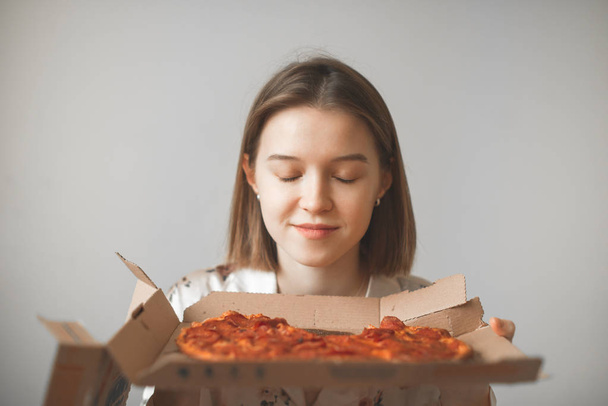 Porträt eines Mädchens hält eine Schachtel heiße Pizza in ihren Händen, riecht den Geruch von Pizza mit geschlossenen Augen, Hintergrund einer hellen Wand.Nettes Mädchen im Schlafanzug mit einer Schachtel Pizza auf grauem Hintergrund - Foto, Bild