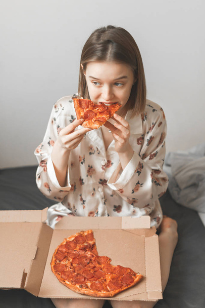 Girl in a pajama sits on the bed in the bedroom with a box on her lap and eats an appetizing piece of pizza. Woman eats a pizza on the bed. Vertical photo. Copyspace - Photo, image