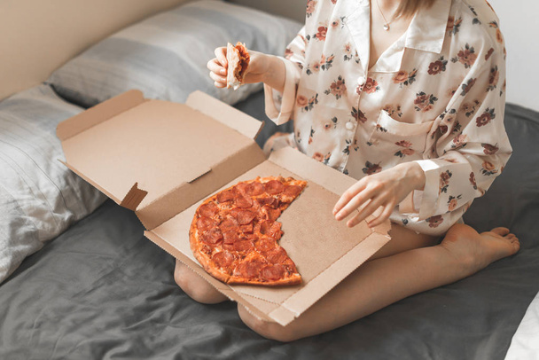 Close up photo, girl in a pajama sitting on a dark sheet, with a box of pizza on her lap, holding a piece of pizza in her hands, eating fast food on the bed. Copyspace - Photo, Image