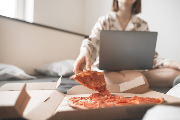 Hungry young girl sits at home on a bed with a pizza box and a laptop, holds a piece of pizza in his hands and is about to eat it. Teen eats at home on a pizza bed with delivery - Foto, Imagen