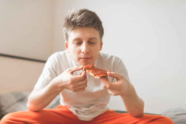 Στενή πορτρέτο ενός άντρα που κάθεται στο κρεβάτι με ένα κομμάτι πίτσα στα χέρια του, τρώει με τα μάτια του κλειστά. Fast Food, πίτσα έννοια. Νεαρός άνδρας τρώει μια πίτσα για πρωινό. - Φωτογραφία, εικόνα