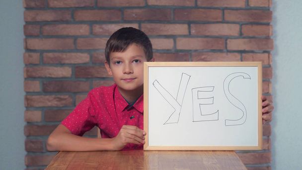Παιδί κάθεται στο γραφείο κρατώντας πινακάκι με γράμματα Ναι o - Φωτογραφία, εικόνα
