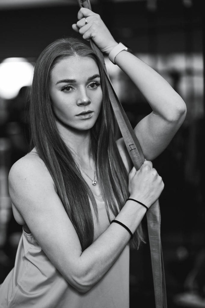 Black and white photo of the beautiful athletic girl dressed in sporty clothes standing in the gym next to the sport equipment - Photo, Image
