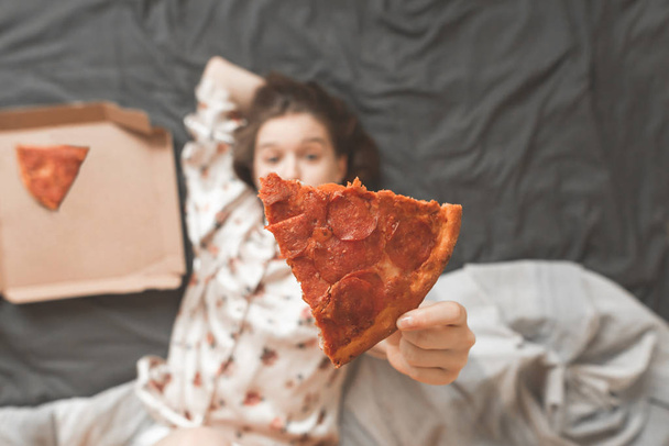 Дівчина в піжамі лежить вдома в ліжку з коробкою для піци, тримаючи шматочок піци в руках і показуючи камеру. Зосередьтеся на шматочку піци, дівчина тримає руки
. - Фото, зображення