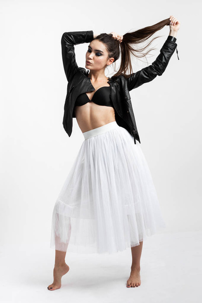 Beautiful brunette girl in a white tulle skirt in a black bra and black leather jacket stands on the white background in the studio - 写真・画像