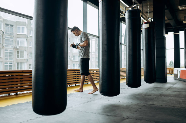 Dark-haired guy dressed in the grey t-shirt and black shorts stands behind the hanging punching bag next to panoramic windows in the boxing gym - Photo, Image