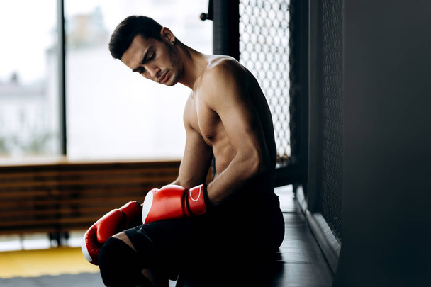 Brutální černovlasý muž s nahý trup a červené Boxerské rukavice na ruce sedí na okraji boxerský ring - Fotografie, Obrázek
