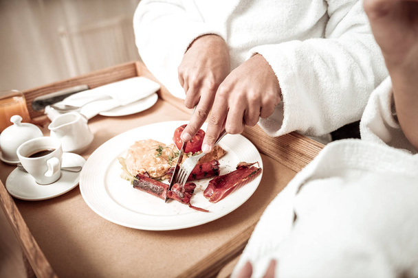 Close up of man wearing bathrobe cutting sausage having breakfast - Foto, Imagen