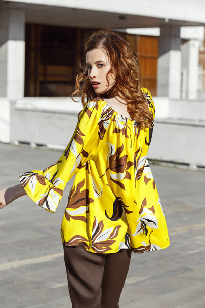 Slim stylish brunette girl dressed in yellow with brown flowers blouse is walking in the street on a sunny day - Foto, afbeelding