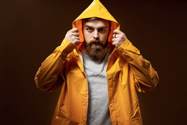 Red-haired guy with a beard and mustache dressed in a gray t-shirt and yellow jacket with a hood is posing on a brown background in the studio - Foto, Imagen
