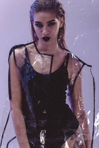 Fashionable model with wet hair and dark lipstick dressed in black swimming suit and transparent rain coat is posing behind the wet  transparent membrane on the white background in the studio - Valokuva, kuva