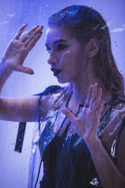 Fashionable model with wet hair dressed in black swimming suit and transparent rain coat is posing behind the wet transparent membrane in the studio with lighting simulating evening street light - Foto, Imagen