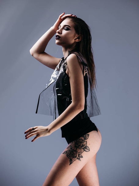Gracile stylish girl with wet hair and tattoo on her leg dressed in black swimming suit and transparent jacket is posing on the gray background in the studio - Foto, imagen