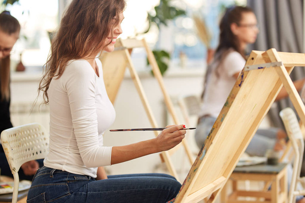 Smiling girl with brown curly hair dressed in white blouse paints a picture at the easel in the drawing school - Photo, image