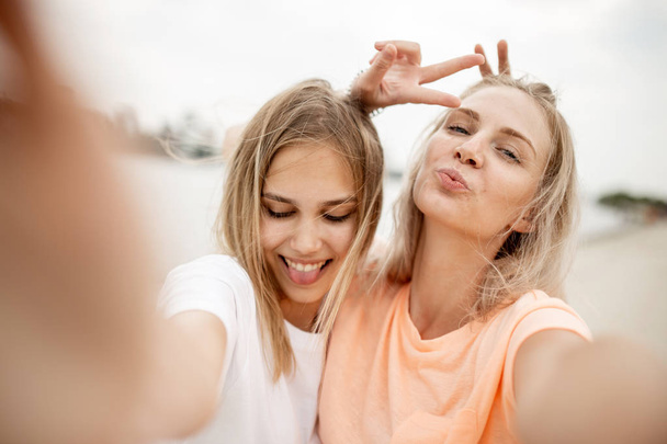 Two young attractive blonde girls take a selfie on the beach on a warm windy day - Foto, Imagem